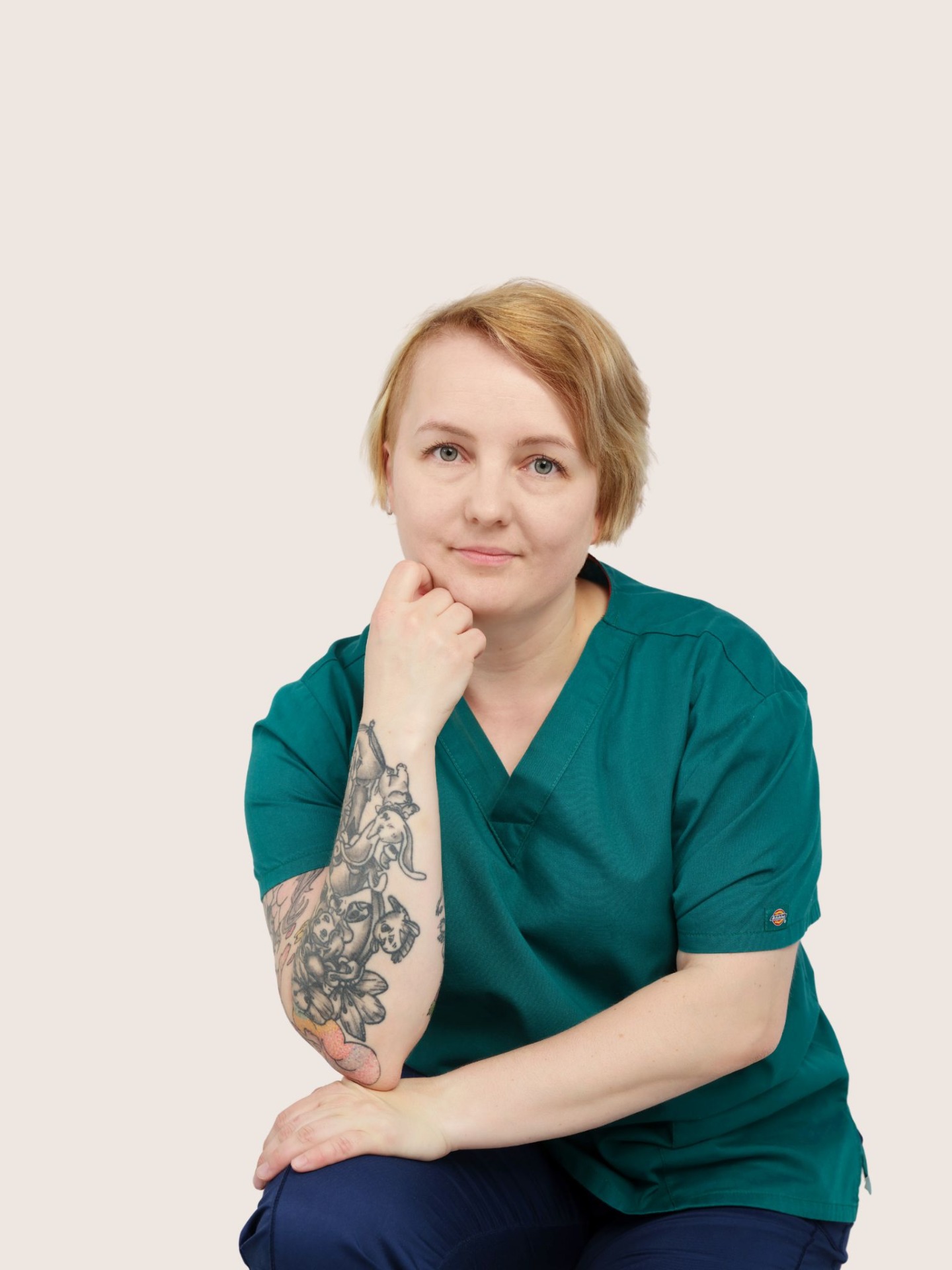 Lyhythiuksinen nainen vihreässä työasussa, käsi leuan alla, hymyilee vaaleaa taustaa vasten.